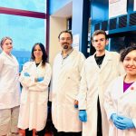 The Zovoilis research team wears labs coats and gloves, smiling in their lab .