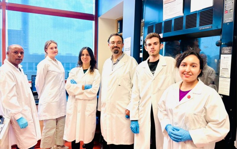 The Zovoilis research team wears labs coats and gloves, smiling in their lab .