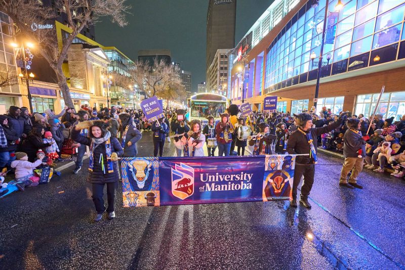 The University of Manitoba contingent participates in Winnipeg's Santa Claus Parade, holding a colorful banner with the university's logo and artwork featuring bison imagery. Participants, bundled up for the cold, wave to the cheering crowds lining the festive, brightly lit downtown street. Signs reading "We Are All Bisons" are visible, along with a brass band adding to the lively atmosphere.