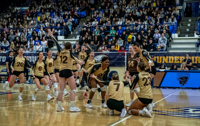 UM Bisons women's volleyball team celebrates on the court after their Canada West championship victory, with cheering players and a packed crowd in the background.