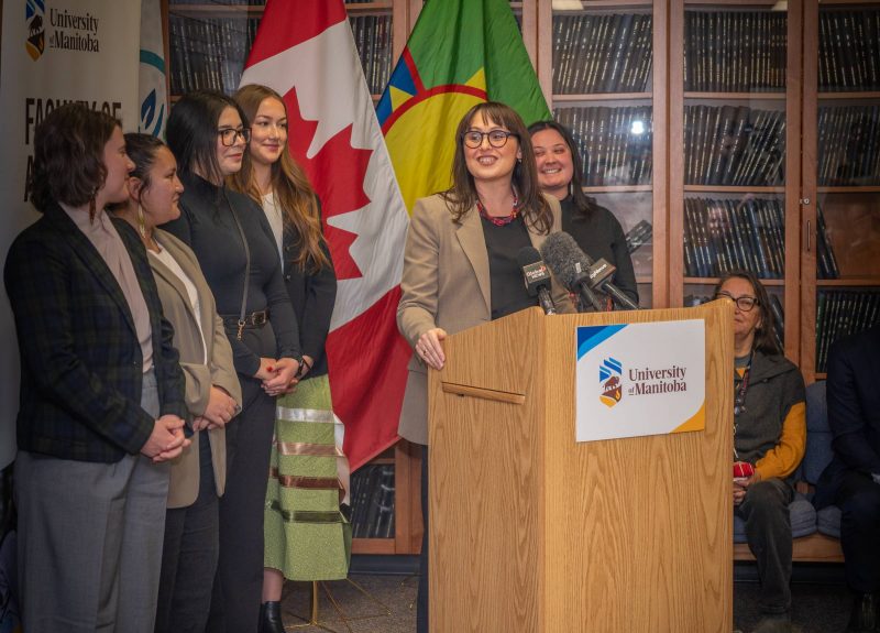 Woman standing at a podium speaking to a crowd. A group of five women stand behind her.