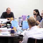 Workshop participants sit around a large table with notebooks and binders.