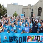 A large group of people gathered outdoors, wearing blue "Bison Pride" T-shirts, holding a banner that reads "Bison Pride" with rainbow-colored letters. They are cheering with their hands raised, showing support for LGBTQ+ pride. The group includes individuals of diverse ages and backgrounds, standing in front of a historic stone building on a sunny day. Some participants hold rainbow flags and UM-branded signs, representing the University of Manitoba's presence at a pride event.