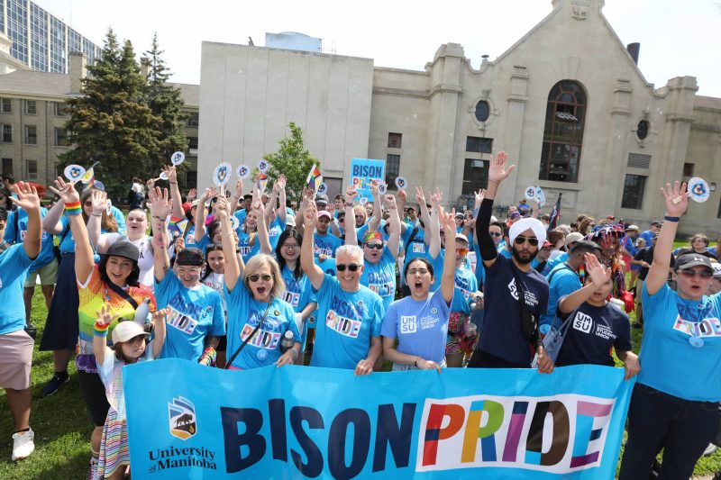 A large group of people gathered outdoors, wearing blue "Bison Pride" T-shirts, holding a banner that reads "Bison Pride" with rainbow-colored letters. They are cheering with their hands raised, showing support for LGBTQ+ pride. The group includes individuals of diverse ages and backgrounds, standing in front of a historic stone building on a sunny day. Some participants hold rainbow flags and UM-branded signs, representing the University of Manitoba's presence at a pride event.