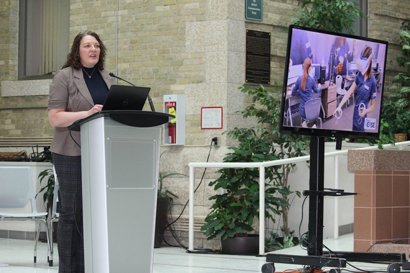 Dr. Nicole Harder stands at a podium. A TV screen shows nursing students wearing VR headsets.