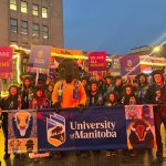 A group of people wearing bison hats with horns, standing with Billy the Bison and holding signs that say "We are all Bisons" and a large University of Manitoba banner.