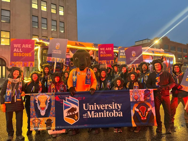 A group of people wearing bison hats with horns, standing with Billy the Bison and holding signs that say "We are all Bisons" and a large University of Manitoba banner.