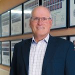 Portrait of Dr. John Perry standing in front of College of Dentistry graduating class photos hanging on a wall.