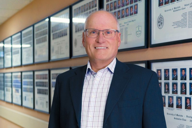 Portrait of Dr. John Perry standing in front of College of Dentistry graduating class photos hanging on a wall.