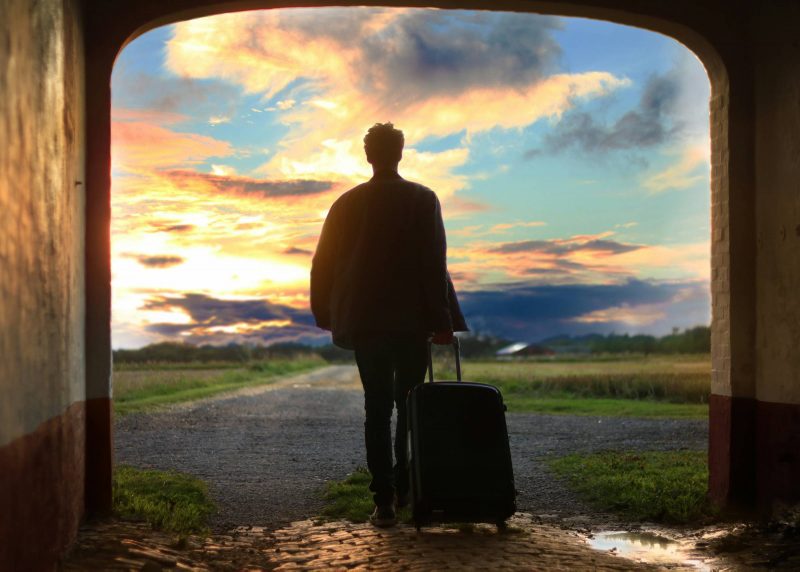 Man pulling hardshell suitcase through a tunnel opening to the runway at sunrise.