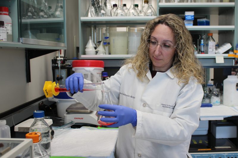 Dr. Inna Rabinovich-Nikitin is in her lab wearing a white lab coat. She is pouring a red liquid from one container to another.