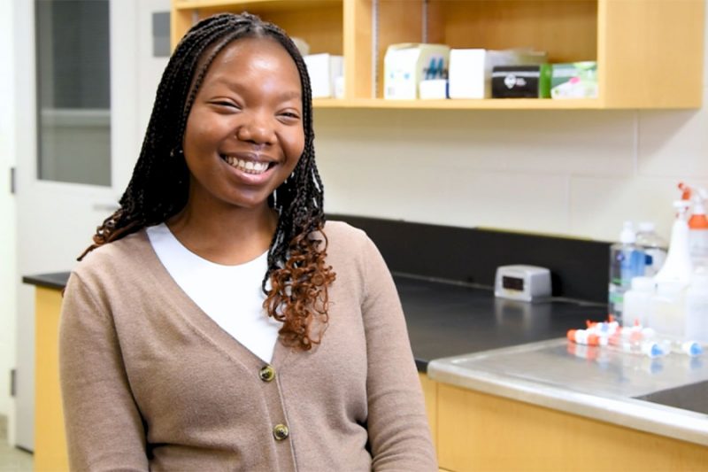 A student smiling at the camera, sitting in a chemistry lab.