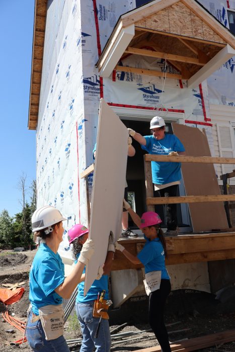 Three people on the ground pass a sheet of construction material to two people standing at the top of a staircase outside. They are all wearing hardhats. 
