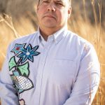 Professor Niigaan Sinclair standing outside in front of a field of tall grasses.