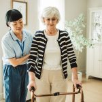 A person helps an older person while they walk using a walker in a house.