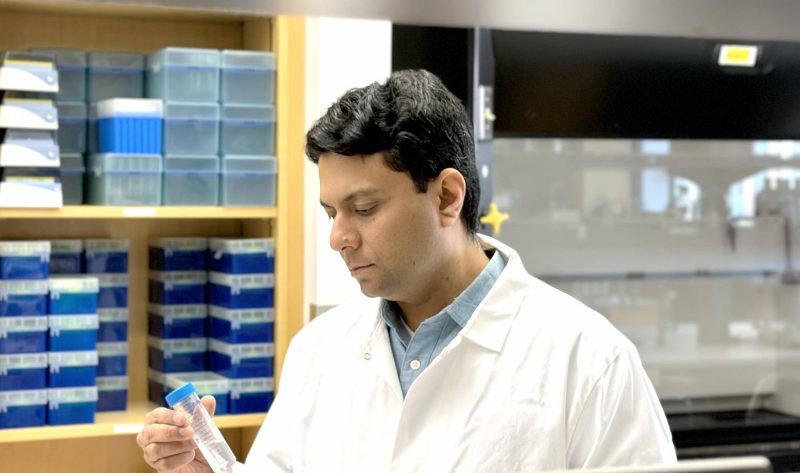 Dr. Abdullah Maruf, wearing a white lab coat, in a laboratory holding a test tube. Shelves with scientific equipment are visible in the background.