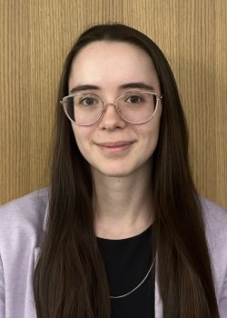 headshot of law student with long brown hair, glasses, black shirt and pink blazer.