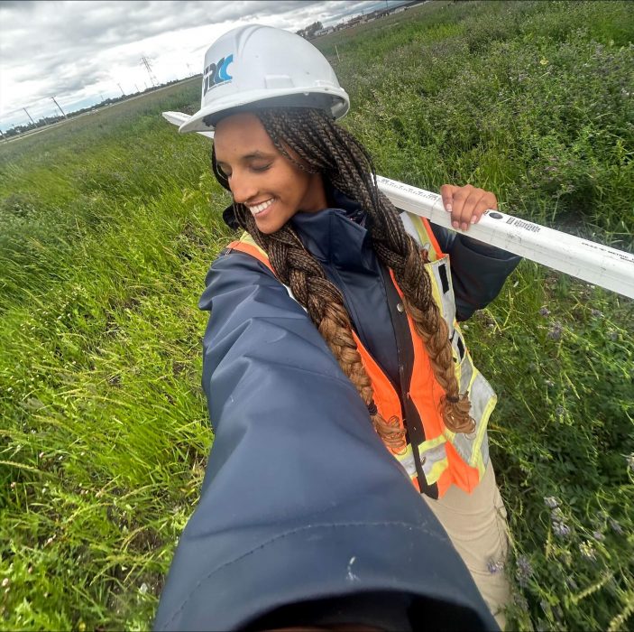 A selfie picture of person wearing a hard hat and safety vest out in a green field