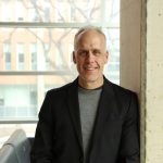 Dr. Jamie Falk smiles at the camera while leaning on a pillar inside the Apotex Centre, with the building’s exterior visible through the windows in the background.