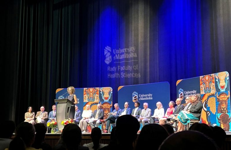 Sixteen people sit in chairs on a stage. Behind them are three banners with UM branding. One person is speaking at a lectern at the front of the stage. The UM and Rady Faculty logo is projected on a curtain.
