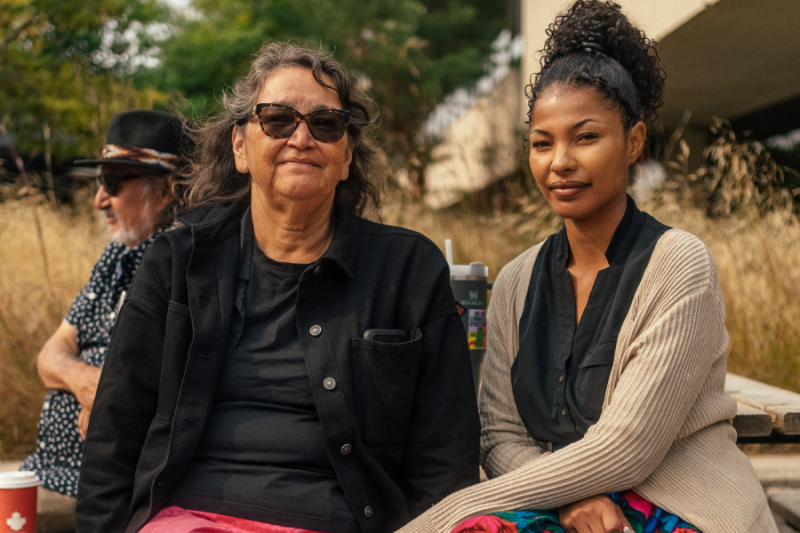 An Indigenous Elder and a young woman sit together outdoors, smiling at the camera during Indigenous Orientation, with another person in the background.