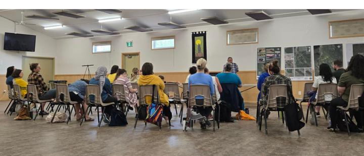 Participants in the Summer Institute sit in a circle during a field trip workshop at Brokenhead, listening to a community speaker at the front of the hall.