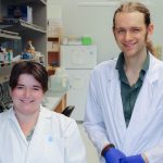 Janessa Sawatzky and Eli Mondor standing side by side in a science lab, smiling at the camera. Lab equipment and shelves are visible in the background.