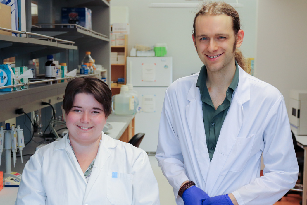 Janessa Sawatzky and Eli Mondor standing side by side in a science lab, smiling at the camera. Lab equipment and shelves are visible in the background.