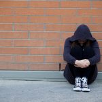 A young person sits on a sidewalk against a wall, head down.