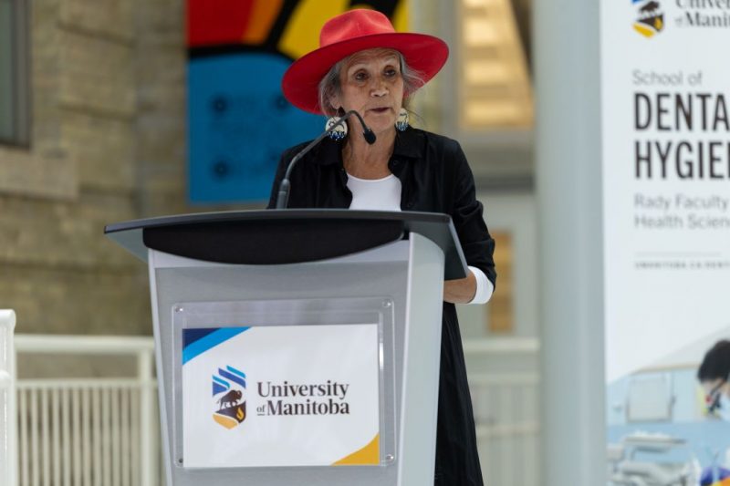 Dr. Mary Jane McCallum speaks into a microphone at a lectern. The UM logo and the words "University of Manitoba" are on the front of the lectern. 