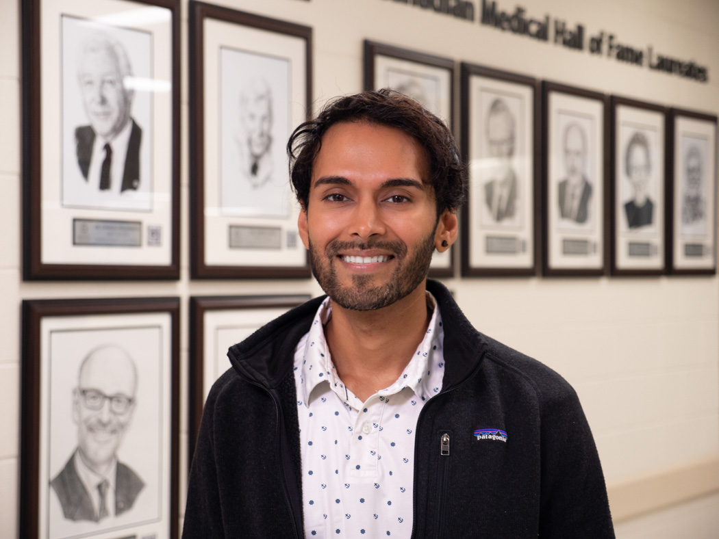 Akhil Ramdoyal smiling in front of the Canadian Medical Hall of Fame Laureates wall at the UM Bannatyne campus.