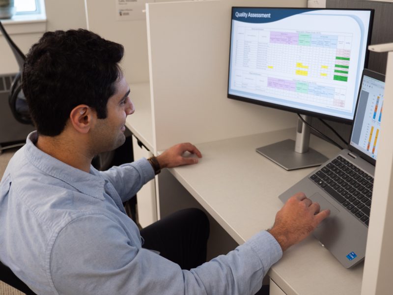 Sam Kavosh sits at a desk, looking at a dual-monitor setup. The left screen displays a table titled 'Quality Assessment'.