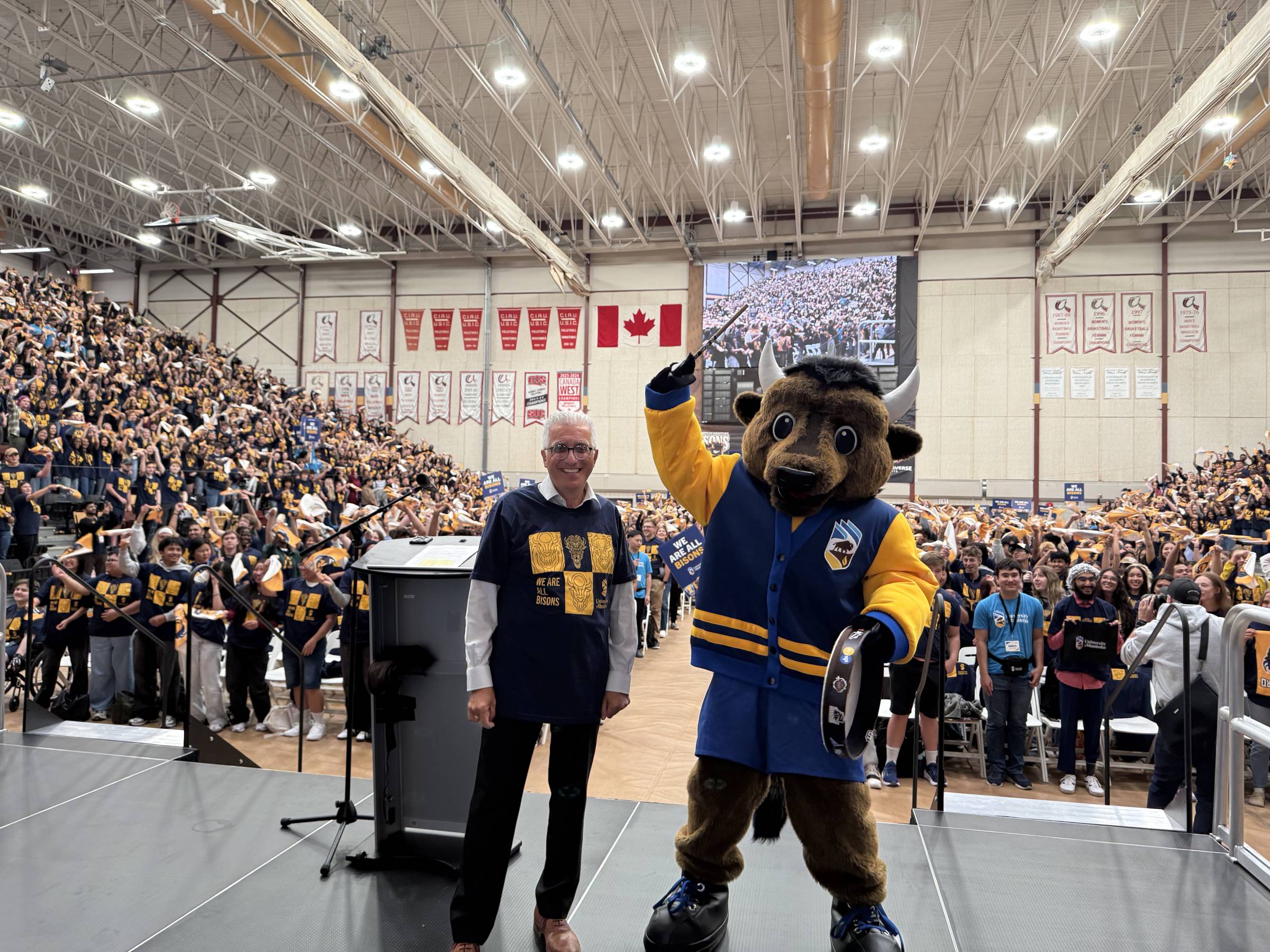 University of Manitoba president and the Bisons mascot stand on stage in front of a packed gymnasium of cheering students during a pep rally. Both wear ‘We Are All Bisons’ shirts, and the crowd holds gold rally towels. A large screen in the background displays the audience.
