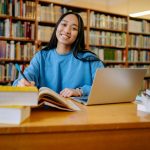 Student in a library with a laptop open and a pile of books open to the left.