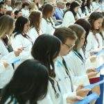 More than 20 students stand in two rows. They are wearing white coats and stethoscopes around their necks. They each hold a program and read the Physician's Pledge.