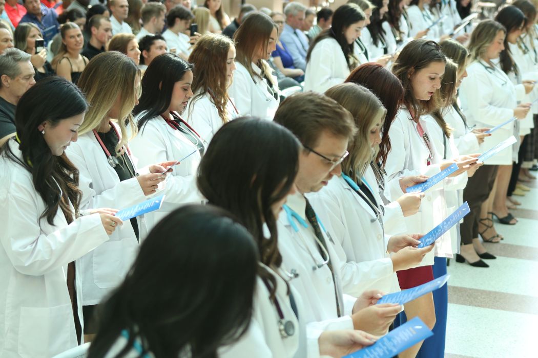 More than 20 students stand in two rows. They are wearing white coats and stethoscopes around their necks. They each hold a program and read the Physician's Pledge.