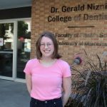 Portrait of Kayla Austin. She is standing outside of a building. On the wall behind her reads "Dr. Gerald Niznick College of Dentistry, Rady Faculty of Health Sciences, University of Manitoba. "
