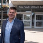 Barret Monchka smiles at the camera while standing outside the Brodie Centre. In the background, the entrance to the Rady Faculty of Health Sciences is visible.