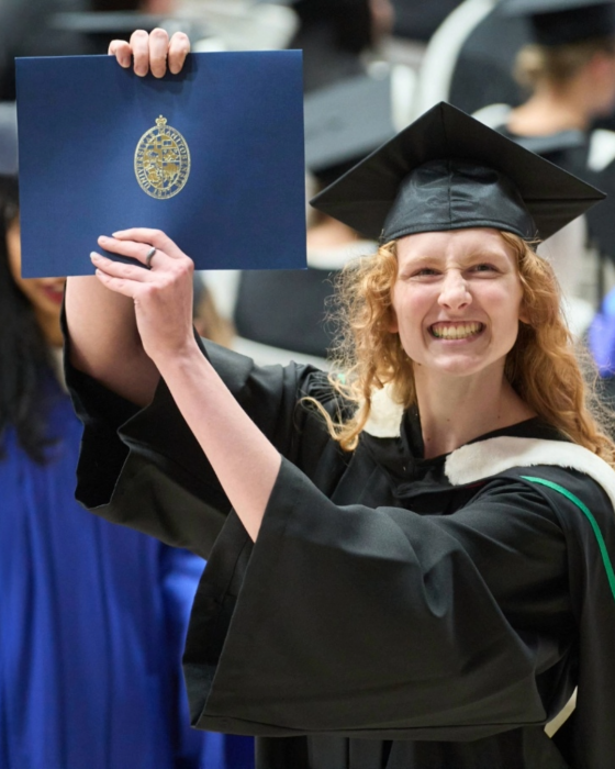 A UM grad wearing a cap and gown holds their parchment in the air with a look of triumph on their face.
