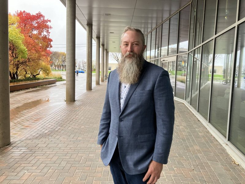 Man in jacket with beard stands outside building