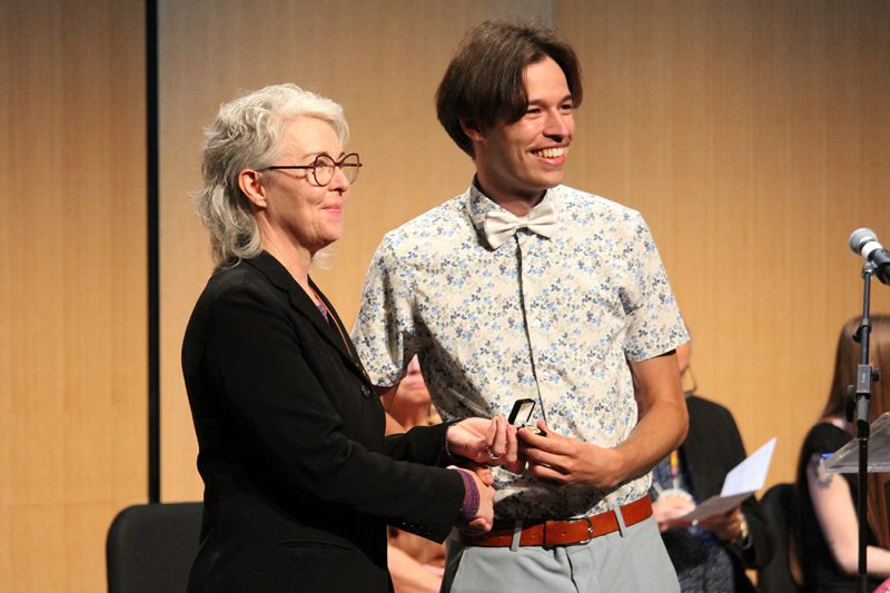 Judah Chepil standing on stage with Colllege of Nursing dean Kellie Thiessen holding his nursing pin.