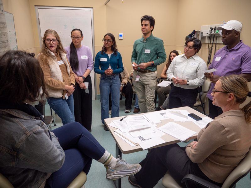 Six students stand while engaging with a Standardized Patient on the left and a facilitator on the right. In the background, other students are seated and observing the interaction.