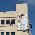 Five people wearing superhero costumes rappel down the side of a building. A large banner with a photo of a child on it reads "The Children's Hospital Foundation of Manitoba. Donate Today! goodbear.ca."