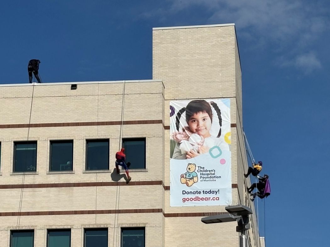 Five people wearing superhero costumes rappel down the side of a building. A large banner with a photo of a child on it reads "The Children's Hospital Foundation of Manitoba. Donate Today! goodbear.ca."