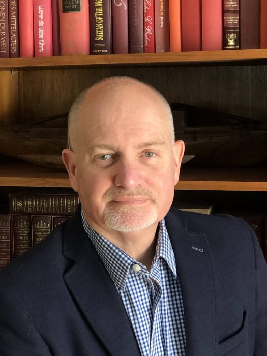 Man in jacket sits before a bookcase