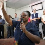 One person holds a microphone and raises their hand. Nine people stand on a stage behind the person with the microphone.