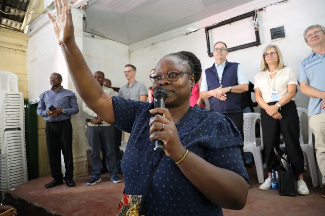 One person holds a microphone and raises their hand. Nine people stand on a stage behind the person with the microphone.