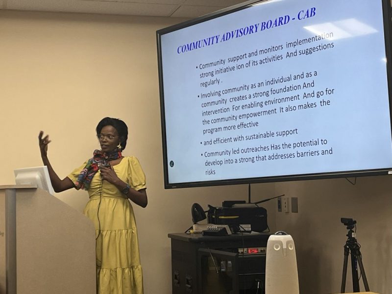Emily Kimemia speaks at a lectern. On a large screen behind her is text with the headline "Community advisory board - CAB."