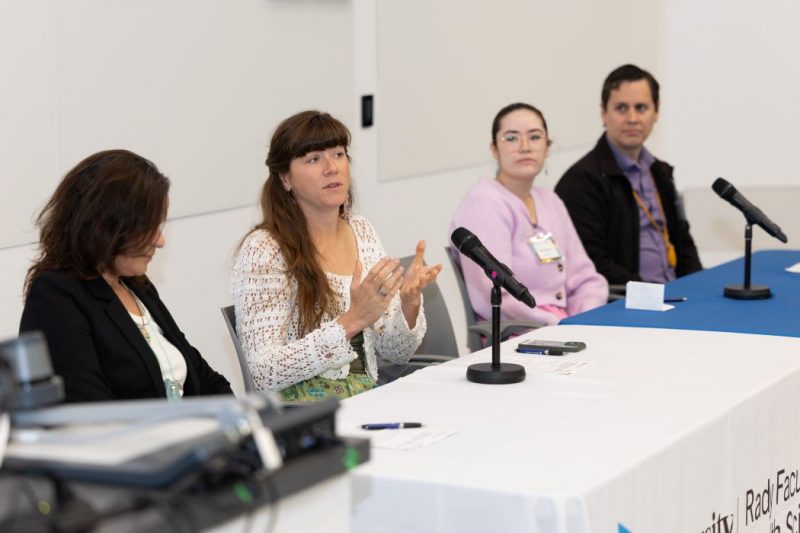 Four symposium panelists are seated at a table as one panelist speaks.