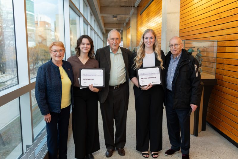 Five people stand and smile at the camera. Two of them are students holding their award certificates.